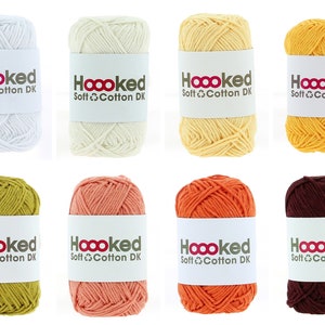 Può includere: Otto gomitoli di filato in vari colori, tra cui bianco, crema, giallo, arancione, rosso, marrone e verde. Ogni gomitolo ha un'etichetta che recita "Hoooked Soft Cotton DK".