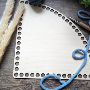 Könnte beinhalten: Eine hellfarbene Holzbasis mit gleichmäßig verteilten Löchern am Rand, entworfen für das Basteln. Eine blaue Kordel ist um die Basis geschlungen, und eine Häkelnadel liegt in der Nähe. Getrocknetes Ziergras sorgt für eine natürliche Note.