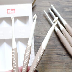 Könnte beinhalten: Ein Set weißer und beigefarbener ergonomischer Häkelnadeln mit einer weißen Box. Die Häkelnadeln haben einen strukturierten beigefarbenen Griff und ein weißes Hakenteil. Die Box zeigt das Markenlogo und die Größen 6,0 und 10,0 mm.