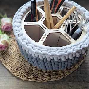 Può includere: Un cestino intrecciato a mano, bicolore grigio e bianco, con un inserto organizer in legno. Il cestino contiene vari strumenti artigianali, tra cui uncinetti e penne. Il cestino è appoggiato su un sottobicchiere intrecciato, con rose rosa sullo sfondo.