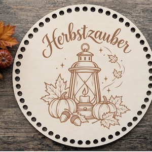 Könnte beinhalten: Eine kreisförmige, helle Holzplatte mit gleichmäßig verteilten Löchern am Rand. In der Mitte befindet sich eine braune Gravur einer Laterne, Kürbissen, Eicheln und Ahornblättern sowie der Schriftzug Herbstzauber darüber.
