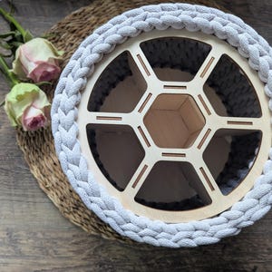 Può includere: Un cesto rotondo intrecciato grigio chiaro con un interno in legno a forma di nido d'ape. Il cesto poggia su un tappetino intrecciato, con due rose rosa e foglie verdi sullo sfondo. Il cesto è un contenitore decorativo.