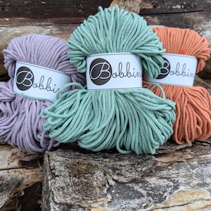 May include: Three spools of cotton rope in different colours: lavender, teal, and orange. Each spool has a white label with the word "Bobbin" printed in black.