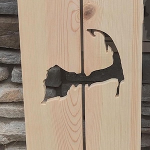 May include: A wooden sign with a cutout of the shape of Cape Cod. The sign is made of two pieces of wood that are joined together. The sign is painted a light brown color.