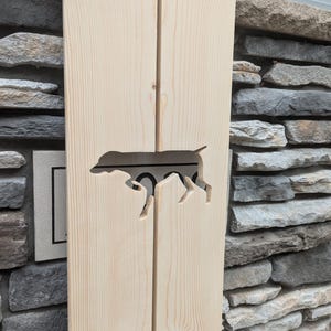 May include: A wooden sign with a cutout of a dog silhouette. The sign is light brown and has a natural wood grain. The dog silhouette is facing to the right.