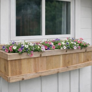 May include: A wooden window box filled with colorful petunias in shades of pink, purple, and white. The rectangular planter is mounted below a white-framed window on a white exterior wall.