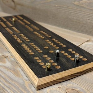 May include: A wooden cribbage board with a black stained surface and a light wood border. The board has brass and silver pegs in the holes.