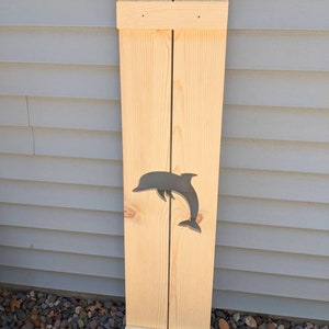 May include: A wooden shutter with a natural finish, featuring a gray dolphin cutout. The shutter is constructed from vertical planks joined by horizontal supports at the top and bottom. The dolphin is centered on the shutter.