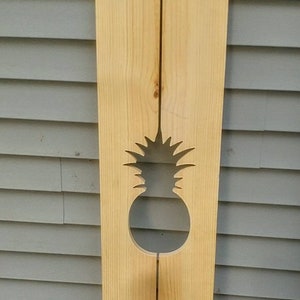 May include: A wooden sign with a cutout pineapple design. The sign is made of two pieces of wood that are joined together. The sign is painted a light brown color.