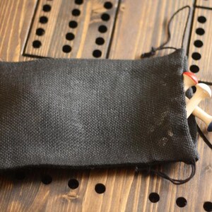 May include: A black burlap bag with a drawstring closure, partially open to reveal wooden pegs with red and blue tops. The bag rests on a wooden surface with drilled holes, likely for a game.