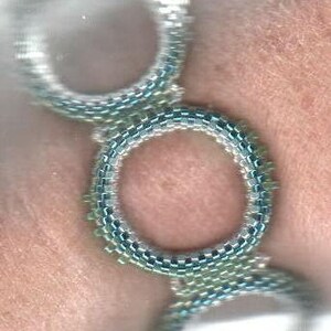 May include: A close-up of a silver and green beaded bracelet with multiple circular designs. The bracelet is worn on a person's wrist.