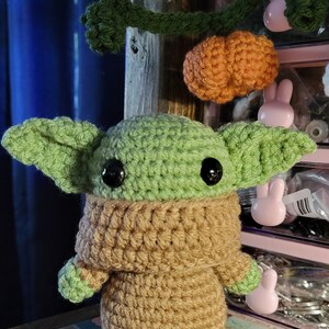 May include: A crocheted green and brown Baby Yoda plush toy with large black eyes. The toy is sitting on a wooden surface with a decorative tile.