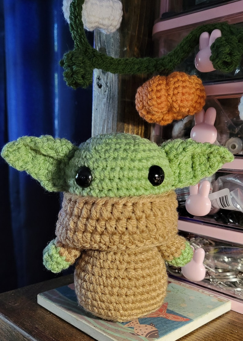 May include: A crocheted green and brown Baby Yoda plush toy with large black eyes. The toy is sitting on a wooden surface with a decorative tile.