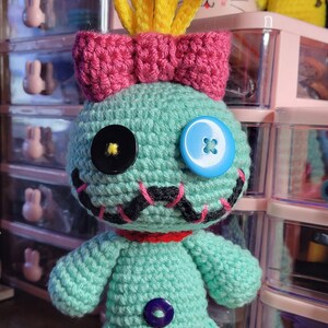 May include: A turquoise crochet doll with a pink bow and yellow hair, resembling a cartoon character. It has button eyes and a stitched mouth. Other crochet toys are visible in the background. The doll is handmade.