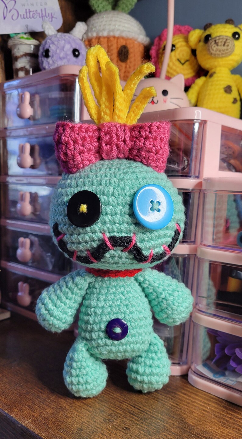May include: A turquoise crochet doll with a pink bow and yellow hair, resembling a cartoon character. It has button eyes and a stitched mouth. Other crochet toys are visible in the background. The doll is handmade.
