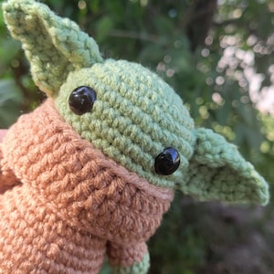 May include: A crocheted plush toy in the shape of a green alien with large black eyes and a brown crocheted hooded robe.