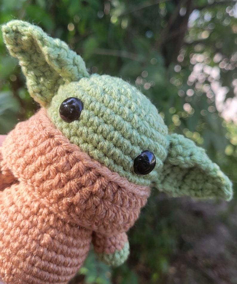 May include: A crocheted plush toy in the shape of a green alien with large black eyes and a brown crocheted hooded robe.