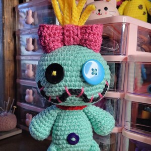 May include: A handmade teal crochet doll with a pink bow, yellow hair, and button eyes. The doll has a stitched mouth and a purple button on its body. Other crochet toys are visible in the background.