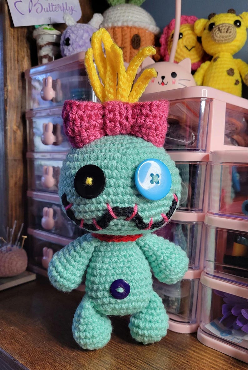 May include: A handmade teal crochet doll with a pink bow, yellow hair, and button eyes. The doll has a stitched mouth and a purple button on its body. Other crochet toys are visible in the background.