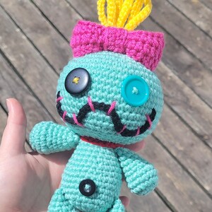 May include: A teal crochet doll with a pink bow and yellow tufts. The doll has button eyes and black stitched details. It has a red collar. The doll is handmade and measures approximately 15 cm in height.