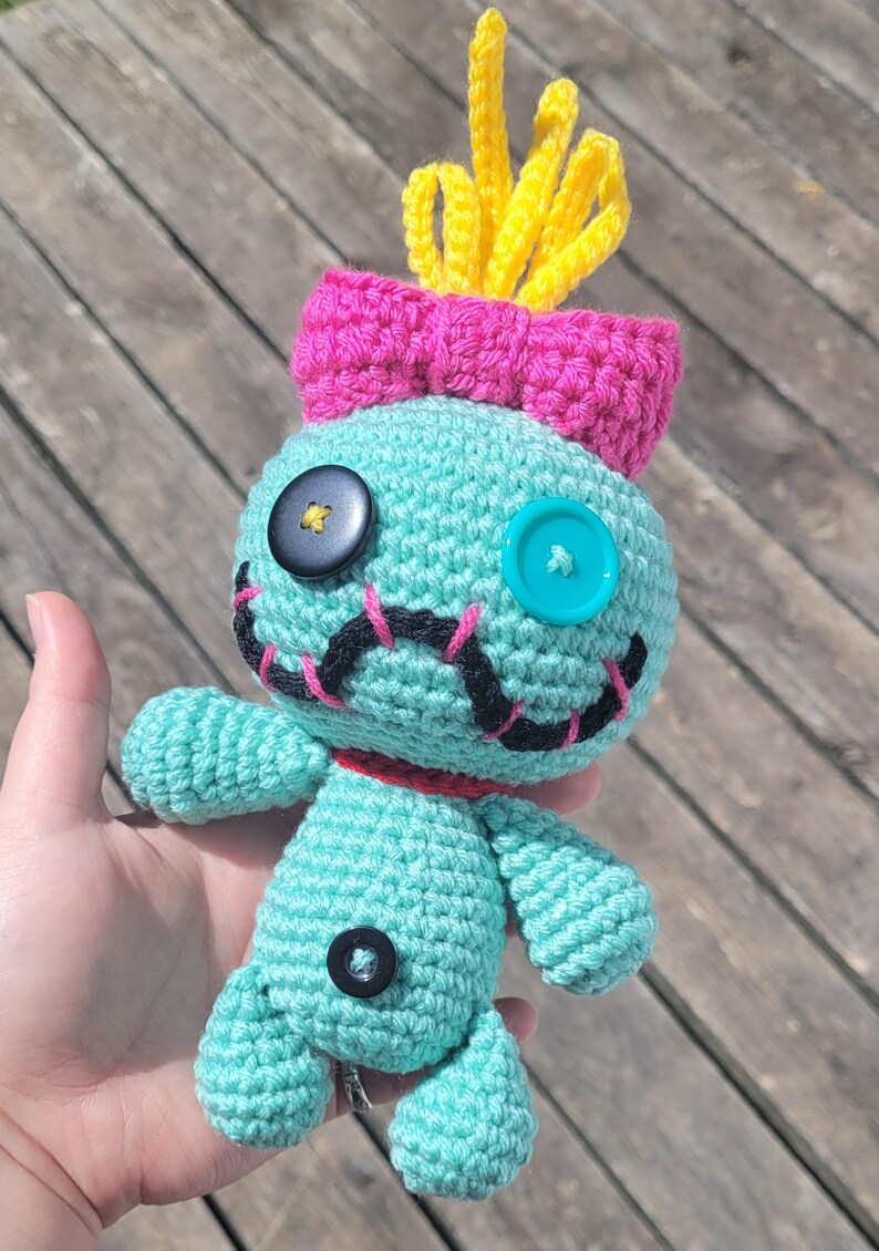 May include: A teal crochet doll with a pink bow and yellow tufts. The doll has button eyes and black stitched details. It has a red collar. The doll is handmade and measures approximately 15 cm in height.