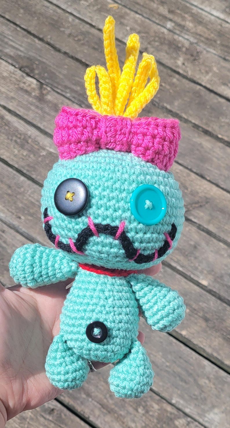 May include: A handmade crochet doll in a light blue colour, featuring a pink bow, yellow hair, and black button eyes. The doll has a stitched smile and a red collar. The doll is approximately 15 cm tall.