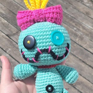 May include: A turquoise crocheted doll with a pink bow, yellow tufts, and button eyes. The doll has black stitched details for the mouth and nose, and a red collar. A unique handmade item.