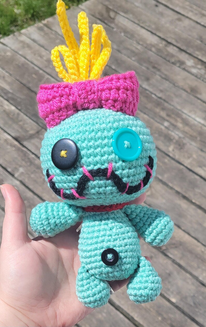 May include: A turquoise crocheted doll with a pink bow, yellow tufts, and button eyes. The doll has black stitched details for the mouth and nose, and a red collar. A unique handmade item.