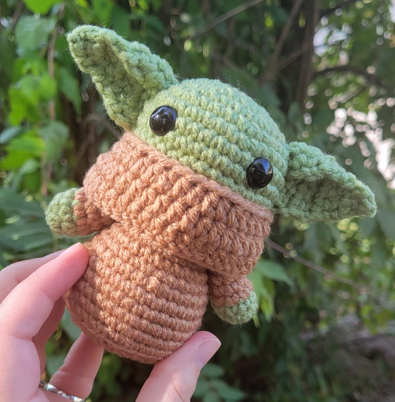 May include: A crocheted plush toy of a green alien character with large eyes and a brown hooded jacket.