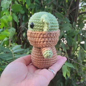 May include: A crocheted green and brown character with a large head and big eyes. The character is wearing a brown hooded robe.