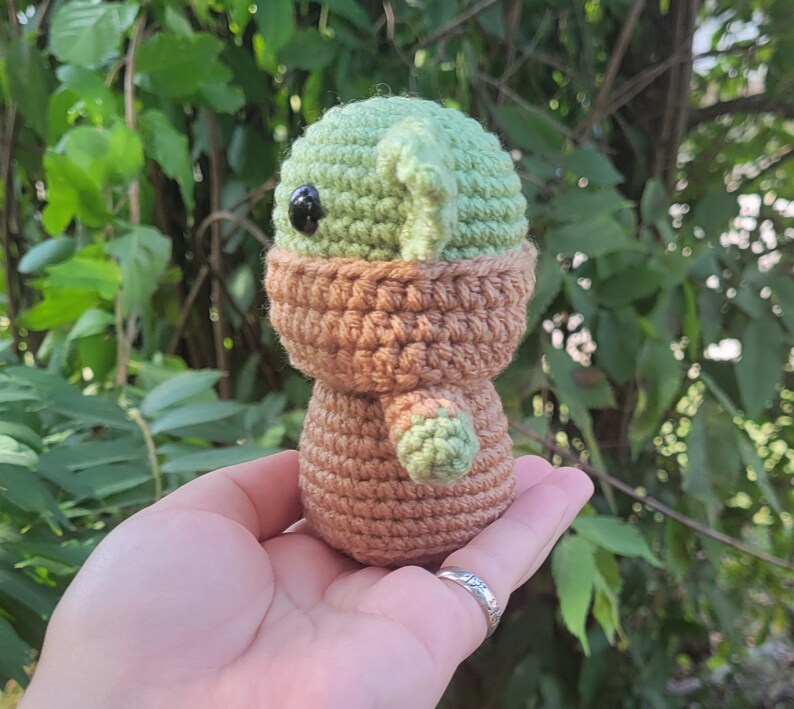 May include: A crocheted green and brown character with a large head and big eyes. The character is wearing a brown hooded robe.