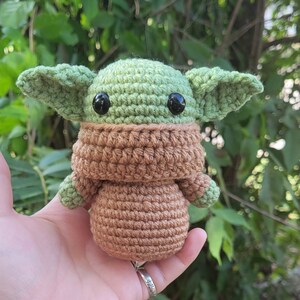 May include: A crocheted green and brown plush toy resembling a character from the Star Wars franchise. The toy has large black eyes and a brown crocheted scarf.