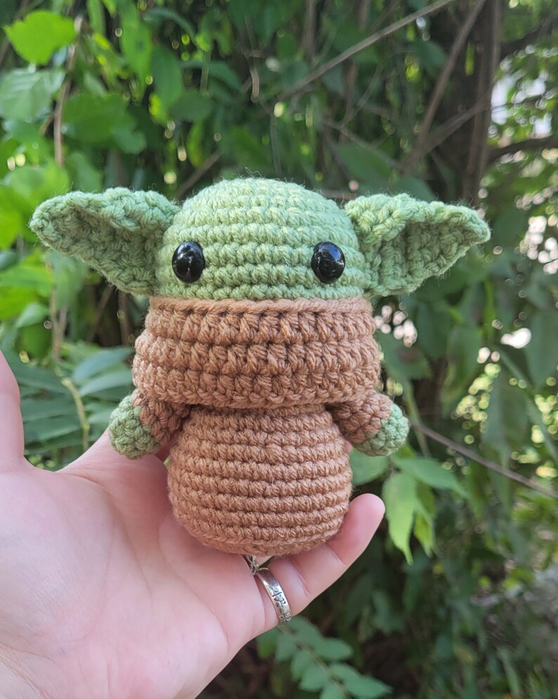 May include: A crocheted green and brown plush toy resembling a character from the Star Wars franchise. The toy has large black eyes and a brown crocheted scarf.