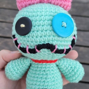 May include: A handmade crochet doll in light teal with a pink bow, black and blue button eyes, and a red collar. The doll has a stitched black mouth with pink accents and yellow yarn hair. The doll is approximately 15 cm tall.