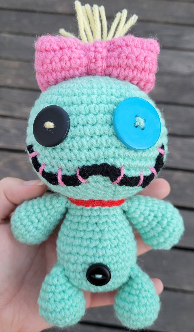 May include: A handmade crochet doll in light teal with a pink bow, black and blue button eyes, and a red collar. The doll has a stitched black mouth with pink accents and yellow yarn hair. The doll is approximately 15 cm tall.
