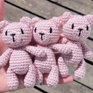 May include: Three pink crocheted teddy bears with black eyes and noses. The bears are sitting on a wooden surface.