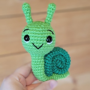 May include: A green crocheted snail with black eyes and a smiling mouth. The snail has a green shell.
