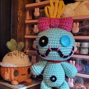 May include: A teal crocheted doll with a pink bow and yellow hair-like features. It has black button eyes, a blue button eye, and a stitched mouth. Next to it is an orange and white crocheted pincushion. The background includes a pink storage unit and other crocheted items.