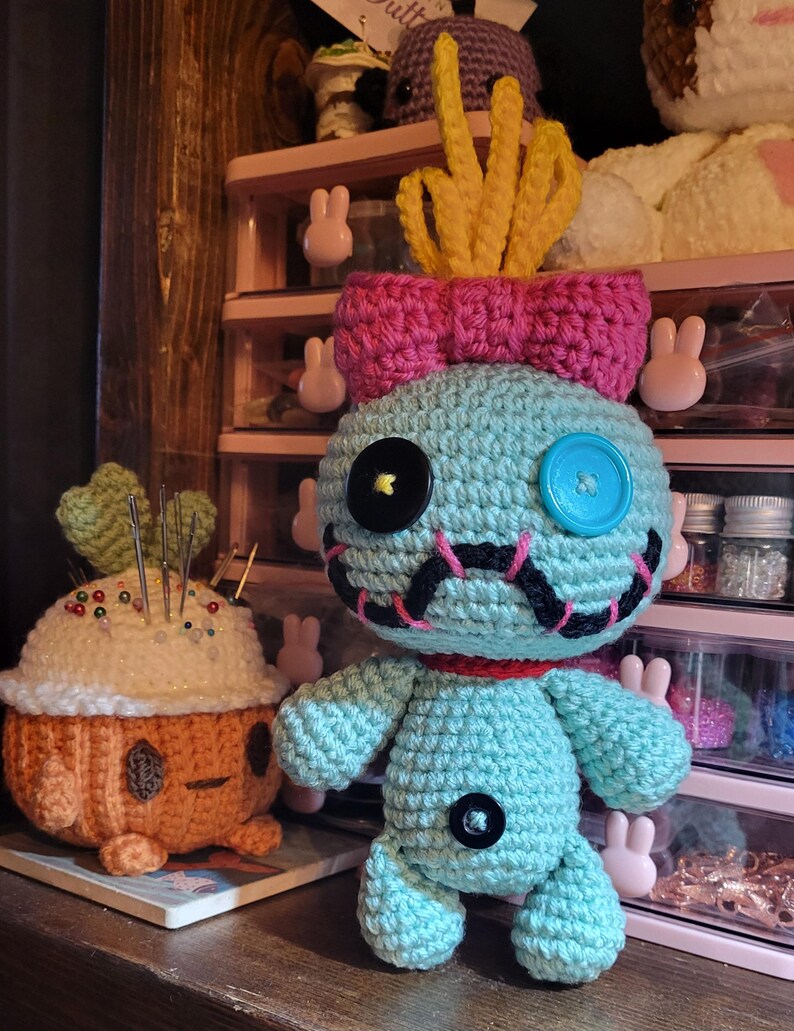 May include: A teal crocheted doll with a pink bow and yellow hair-like features. It has black button eyes, a blue button eye, and a stitched mouth. Next to it is an orange and white crocheted pincushion. The background includes a pink storage unit and other crocheted items.