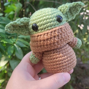 May include: A crocheted green and brown baby Yoda doll with black eyes. The doll is wearing a brown crocheted coat.