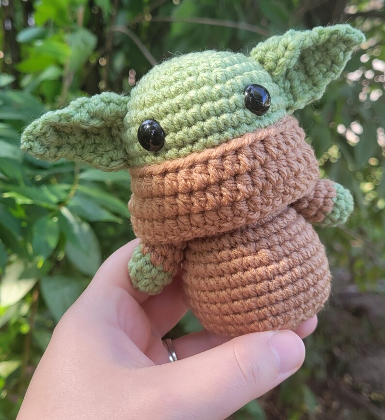 May include: A crocheted green and brown baby Yoda doll with black eyes. The doll is wearing a brown crocheted coat.