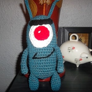 Könnte beinhalten: Ein gehäkeltes Plüschtier von Plankton aus der Zeichentrickserie SpongeBob Schwammkopf. Das Spielzeug ist blau mit einem großen weißen Auge mit einer roten Pupille und einem schwarzen Augenklappen. Das Spielzeug hat ein schwarzes Lächeln und zwei lange Antennen.