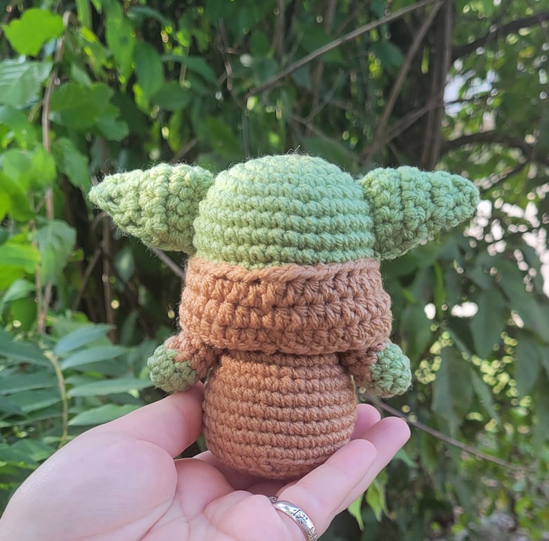 May include: A crocheted green and brown Yoda doll. The doll is holding its hands in front of it and has a brown crocheted robe.