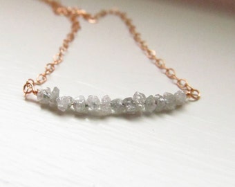 May include: A delicate necklace featuring a rose gold chain and a row of rough, gray-white diamond crystals. The crystals are irregularly shaped, adding a natural, organic feel to the jewelry. The necklace is displayed on a white surface.
