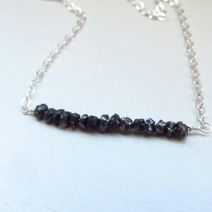 May include: A silver chain necklace featuring a bar of rough, dark gray gemstones. The gemstones are clustered together, creating a textured appearance. The chain is delicate and the necklace is set against a light background.