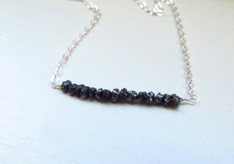 May include: A silver chain necklace featuring a bar of rough, dark gray gemstones. The gemstones are clustered together, creating a textured appearance. The chain is delicate and the necklace is set against a light background.