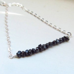 May include: A silver necklace featuring a row of dark, rough-cut gemstones. The gemstones are strung horizontally across the center of the necklace, with a delicate silver chain extending on either side. The necklace is displayed against a light background.