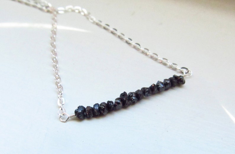 May include: A silver necklace featuring a row of dark, rough-cut gemstones. The gemstones are strung horizontally across the center of the necklace, with a delicate silver chain extending on either side. The necklace is displayed against a light background.