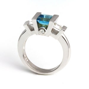 May include: A silver ring featuring a blue topaz centre stone and two smaller diamond side stones.
