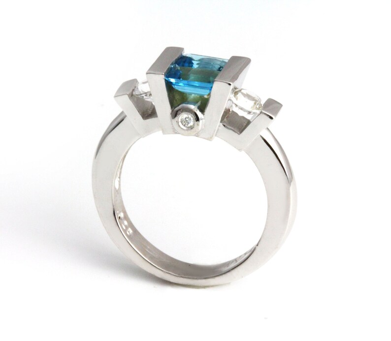 May include: A silver ring featuring a blue topaz centre stone and two smaller diamond side stones.